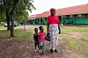 Kvinne med to barn står foran en grønn skolebygning på landsbygda.