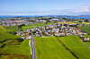 Luftfoto av et tettsted med veier, hus og store grønne jorder under klar himmel.