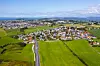 Luftfoto av et tettsted med veier, hus og store grønne jorder under klar himmel.