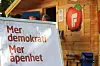 Skilt med teksten «Mer demokrati» og «Mer åpenhet» foran en trebygning med rødt logo-skilt.