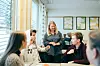 Flere personer sitter rundt et bord i et lyst kontor og snakker med en kvinne stående ved enden.
