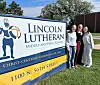 Skilt for Lincoln Lutheran med tre personer stående ved siden av på gresset.