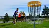 Person kaster en disk mot en disc golf-kurv på en grønn bane med tilskuere i bakgrunnen.