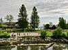 Folk ved en sandstrand og lekeplass langs vannet med trær, hus og overskyet himmel.