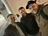 Tre menn smiler i en innendørs selfie, tatt i skrå vinkel.