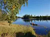 To personer padler kano på en rolig innsjø med gress, trær og skog i bakgrunnen.
