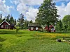 Rød hytte i et grønt landskap med trær, gress og blå himmel med skyer.