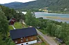 Panoramautsikt over elv, grønn dal, skog og spredte hus ved en grusvei.