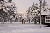 Snødekt park med trær, lyktestolpe og bygninger foran fjell i vinterlandskap.