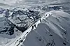 Flere skikjørere på en smal snødekt fjellrygg med høye fjell rundt.