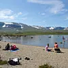 Folk sitter og bader ved en steinstrand med fjell og snødekte topper i bakgrunnen.