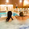 To personer i boblebad ved innendørs basseng, mens en person står på kanten med armene i været.