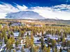 Snødekt fjell over en skogkledd dal med høstfarger og noen små hytter.