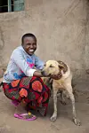 Smilende jente i Guje i Tanzania sitter ved siden av en hund foran en slitt vegg.