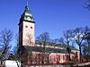Rød murkirke med klokketårn mot blå himmel og nakne trær.
