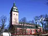 Rød murkirke med klokketårn mot blå himmel og nakne trær.