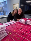 To personer står ved et bord fullt av rosa bøker i et kontor med vindu.