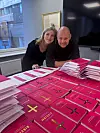To personer står ved et bord fullt av rosa bøker i et kontor med vindu.