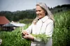 Kvinne i lys jakke holder opp grønne planter i et jordbrukslandskap.