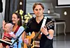 Mann spiller gitar og synger på en liten scene, med kvinne og barn i bakgrunnen.