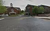 To bygninger ved en asfaltert parkeringsplass med trær og overskyet himmel.