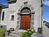 Steinkirke med utsmykket tredør, blomsterbed og gate i sollys.