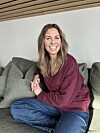 Smilende kvinne i burgunder genser sitter i en grå sofa.
