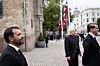 Menn i dresser går gjennom en brosteinsbelagt bygate med flagg og historiske bygninger.