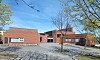 Lav rødbrun kirkebygning ved en asfaltert plass, omgitt av trær under blå himmel.