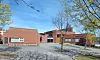 Lav rødbrun kirkebygning ved en asfaltert plass, omgitt av trær under blå himmel.