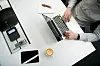 Person skriver på tastatur ved et hvitt kontorpult med telefon, nettbrett og kaffekopp.
