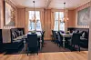 Elegant restaurant med mørke stoler, spisebord, sofaer, store vinduer og klassisk interiør.