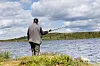 Person fisker ved en innsjø med fjell, lav vegetasjon og skyer i bakgrunnen.